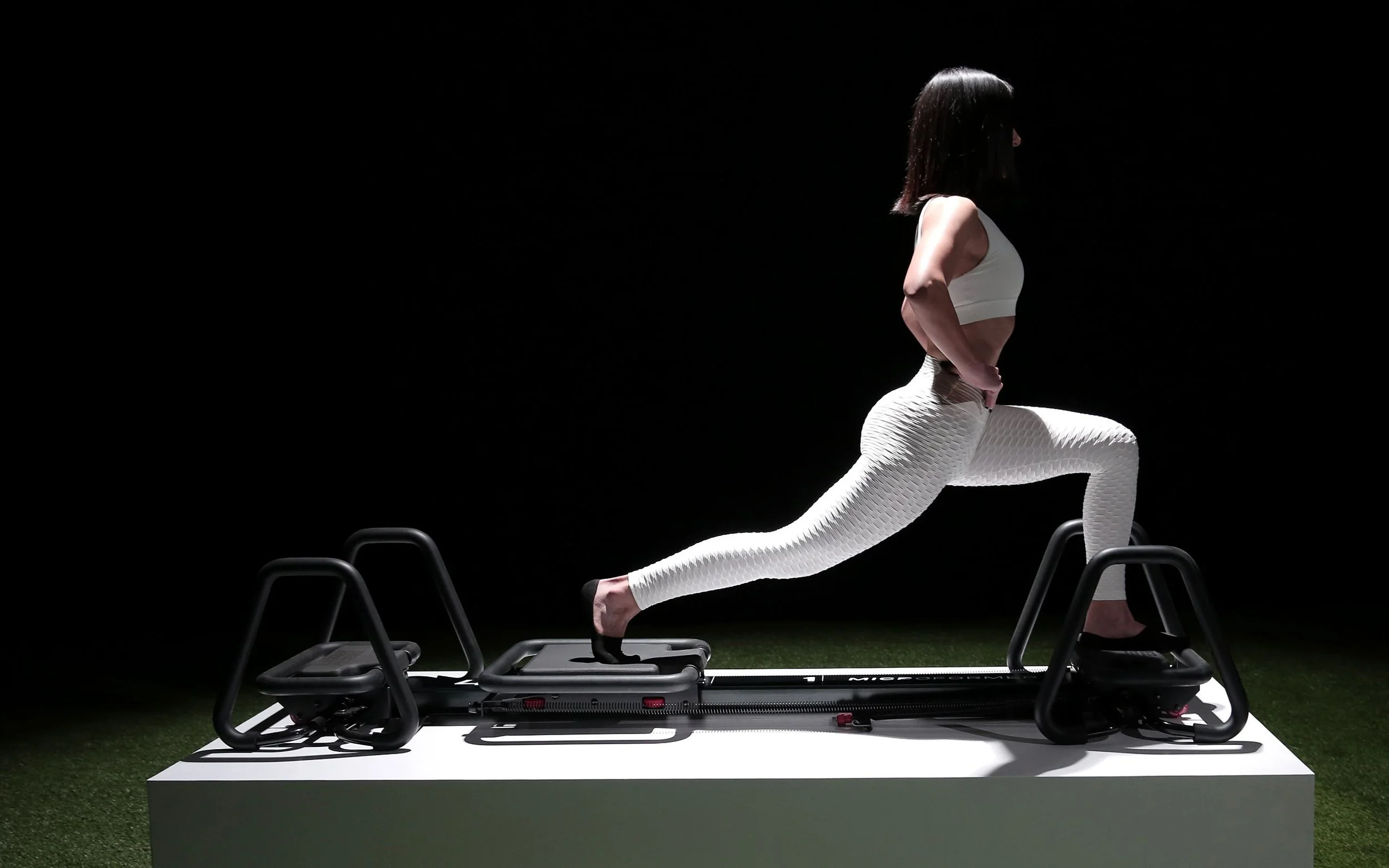 A woman in white workout clothes is doing yoga on a step platform, with her hands on her hips, in a dark studio with chairs around her.
