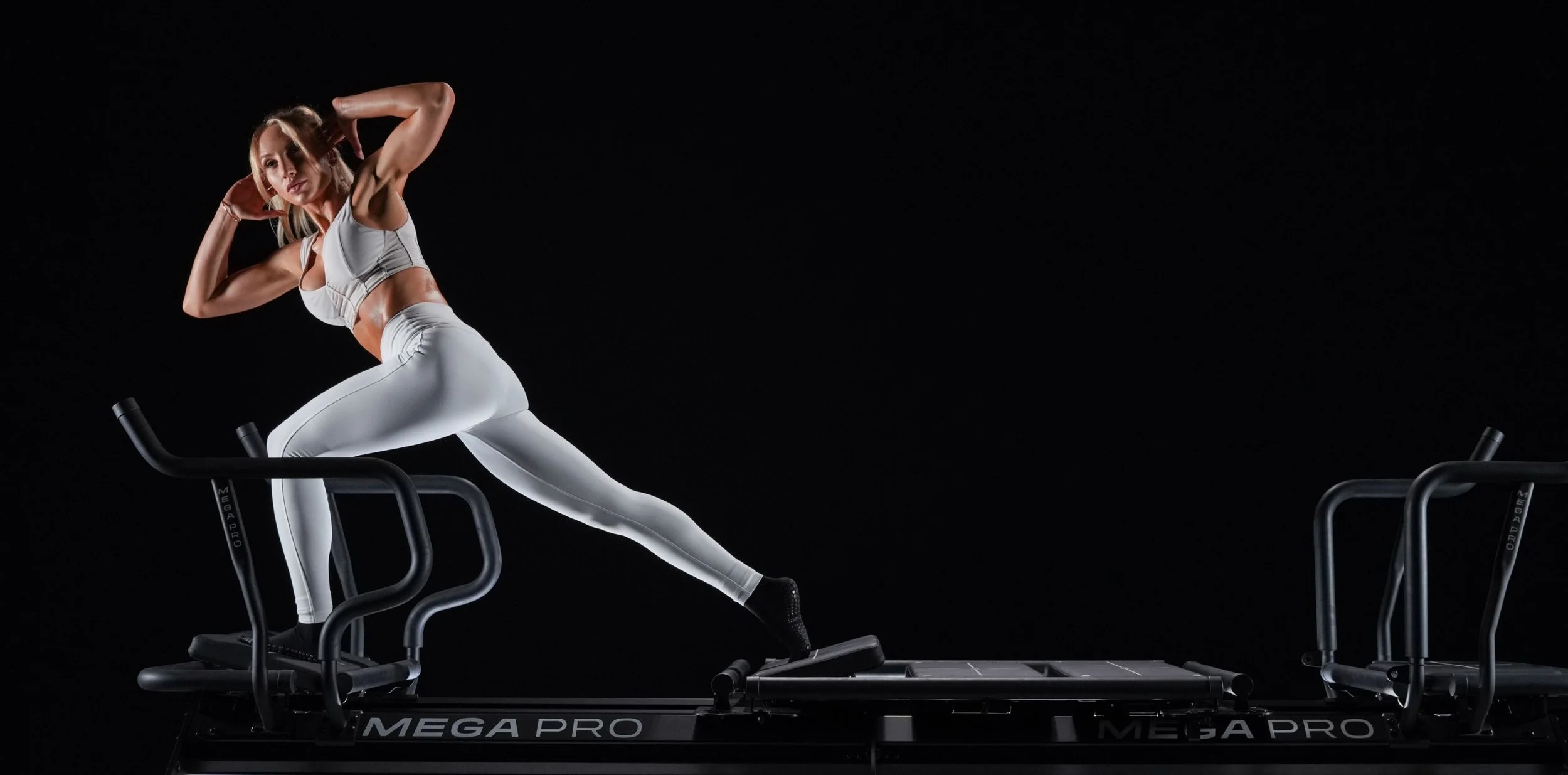A woman in athletic wear performs a stretching pose on a piece of exercise equipment against a black background.