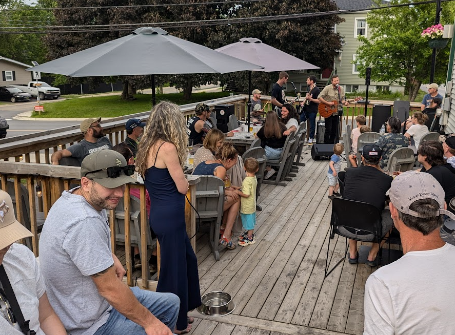 A group of people gathered on a wooden deck outdoors, watching a live musical performance by a band playing guitars.