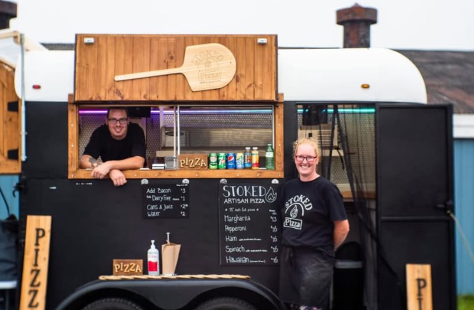 Two smiling women operating a black mobile pizza cart with a wooden sign and a menu board outside during daytime.