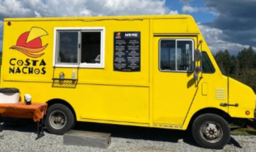 A yellow food truck with a logo of a nacho chip with a red hat and the text "Costa Nachos" on the side. The truck has a window for ordering, a menu board, and is parked outdoors on a gravel surface with a cloudy sky and trees in the background.