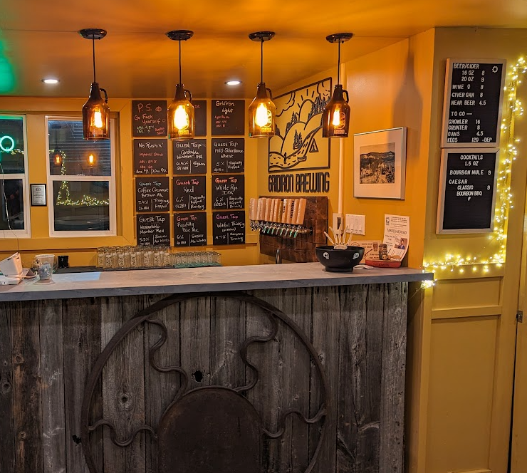 Photo of a bar counter inside a restaurant, with a blackboard menu, beer taps, and decorative lights. The bar front is made of wood with a mounted antlered deer head emblem.