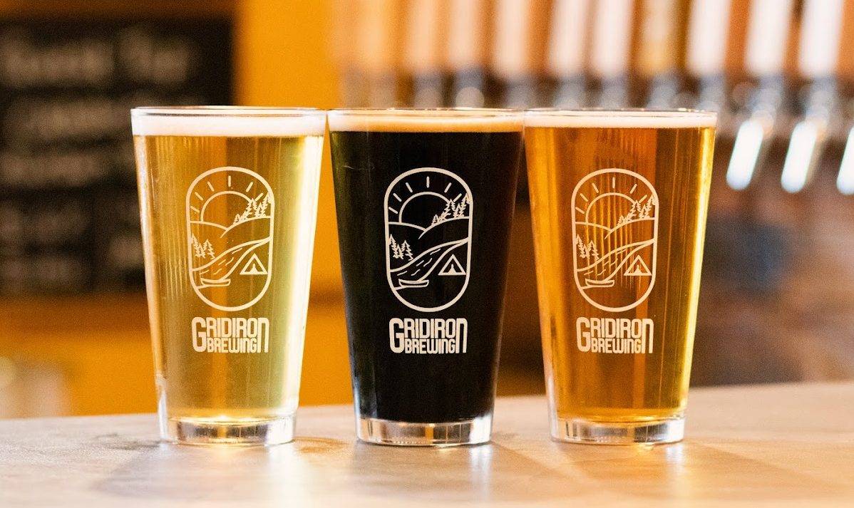 Three glasses of beer with the Glacier Brewing logo on a wooden bar counter, with blurred background.