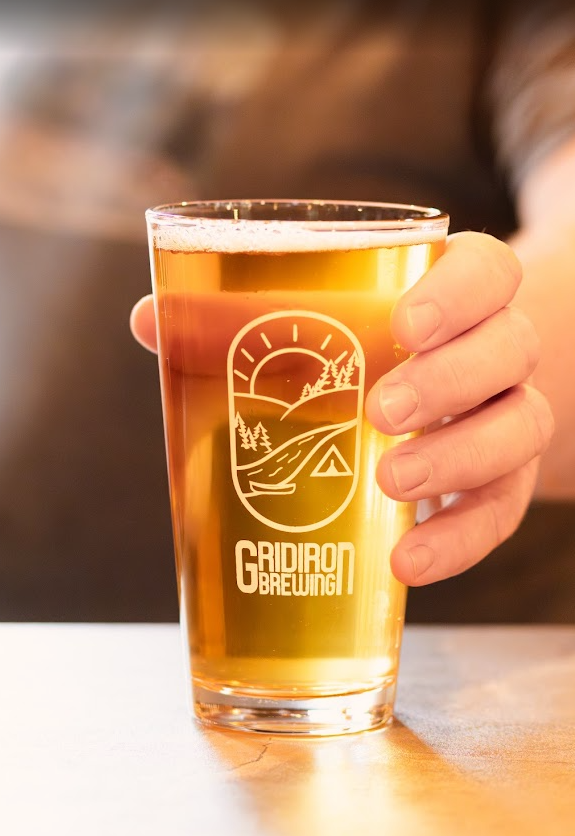 A hand holding a pint glass filled with amber beer, with a logo from Gidrod Brewing on the glass featuring a sunset, mountains, a lake, and a tent.