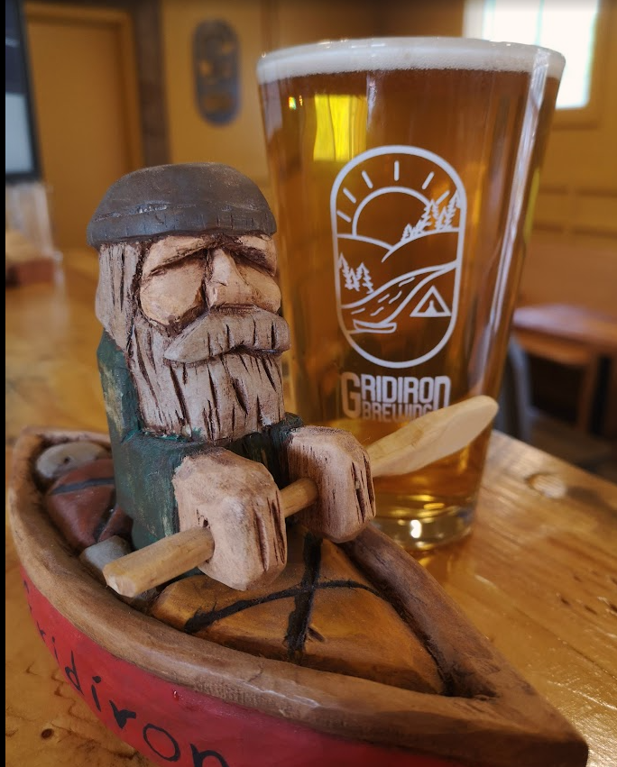 A wooden figurine of a bearded man wearing a cap, sitting in a small boat, holding a paddle. Next to it is a glass of beer with a logo from Gridiron Brewing, on a wooden table inside a cozy, wood-paneled room.
