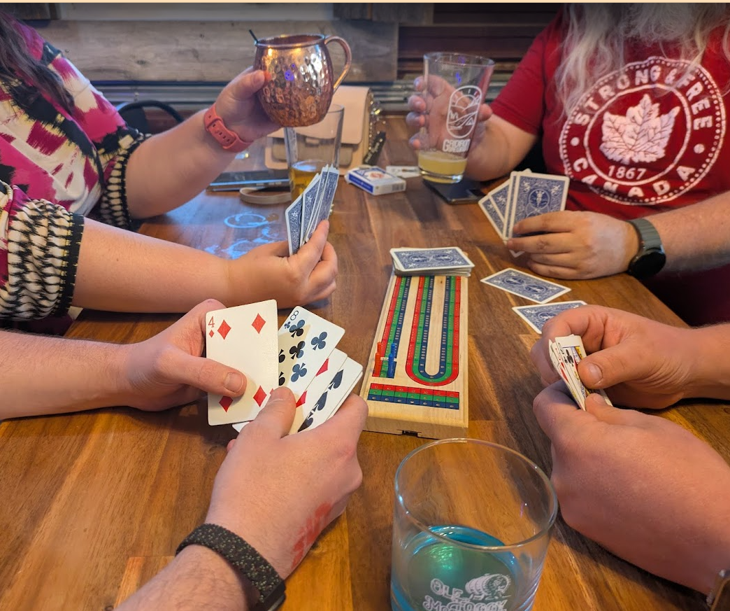 Four people playing a card game at a wooden table. One person is holding a handful of playing cards, with a Royal Flush of hearts visible. There is a colorful vintage game piece track on the table, along with several glasses of beer and a copper mug.
