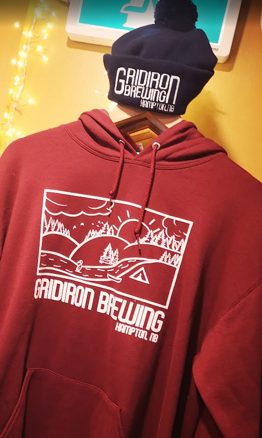 A red hoodie with a white graphic of a landscape with trees, mountains, a river, and sails, with the text "Gridiron Bowning Hampton, NB" printed on it, hanging on a hanger. A black beanie with the same text embroidered on it is placed above it. The b