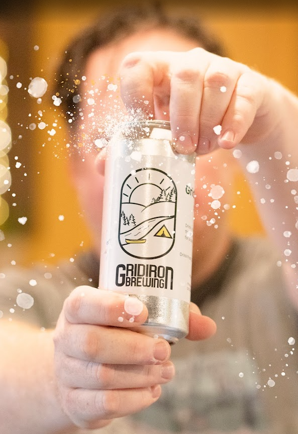 Person opening a can of beverage labeled 'Gridiron Brewing', with foam or spray splashing out.