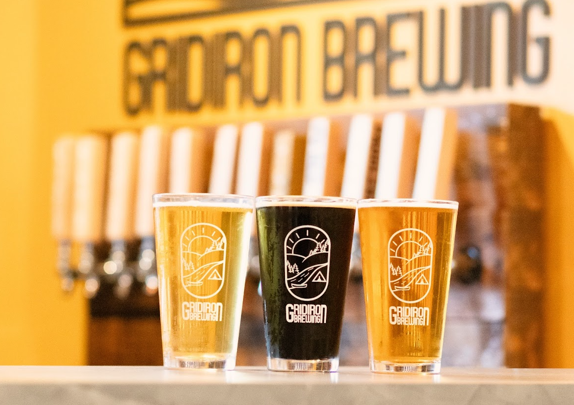 Three glasses of beer on a table, with the logo of Gridiron Brewing on each glass, featuring a stylized landscape with the sun, trees, and a tent, in front of a bar or brewery backdrop.