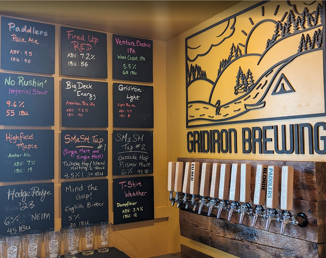 Wall of black chalkboards with beer descriptions and ratings, a row of beer taps labeled with beer names, and a large outdoor-themed logo reading 'Gridiron Brewing' on the wall.