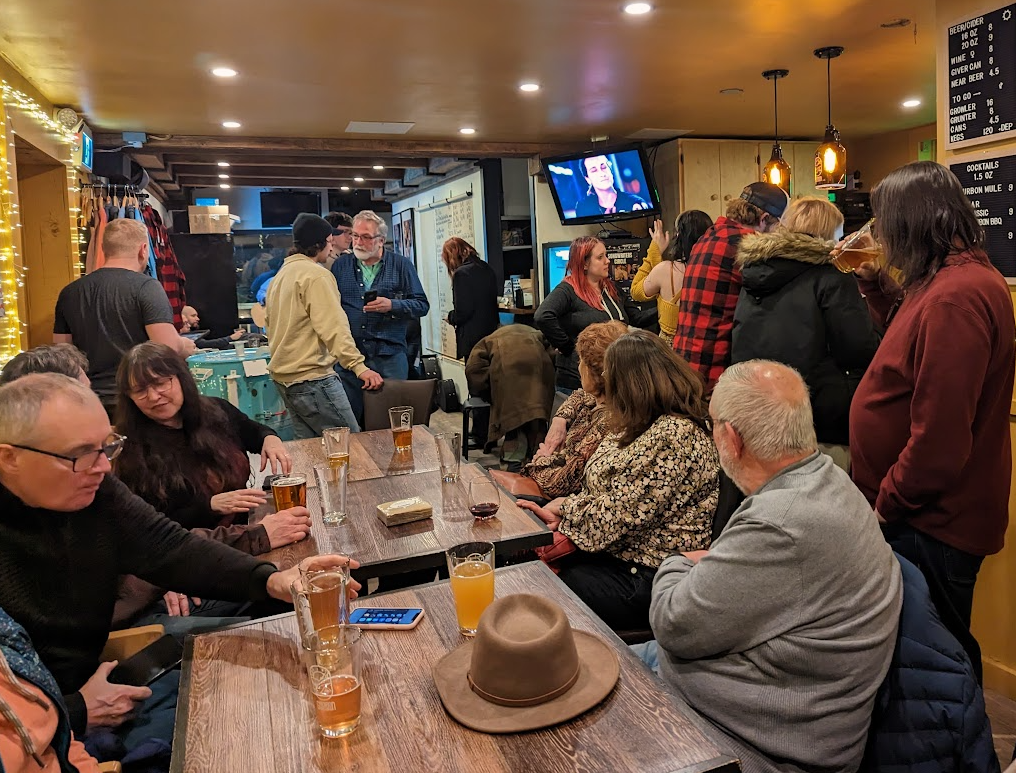 People gathering in a lively bar, sitting and standing around tables with drinks, with a menu board on the wall and a TV showing a person. Cozy lighting and casual atmosphere.