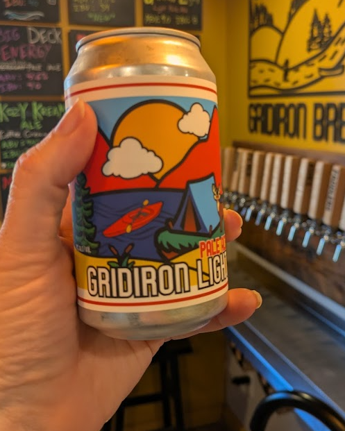 A hand holding a can with colorful outdoor-themed artwork featuring a sun, clouds, a lake, a tent, and a kayak, with the text 'Gridiron Light' and 'Pale Ale' on the label, in a brewery setting.