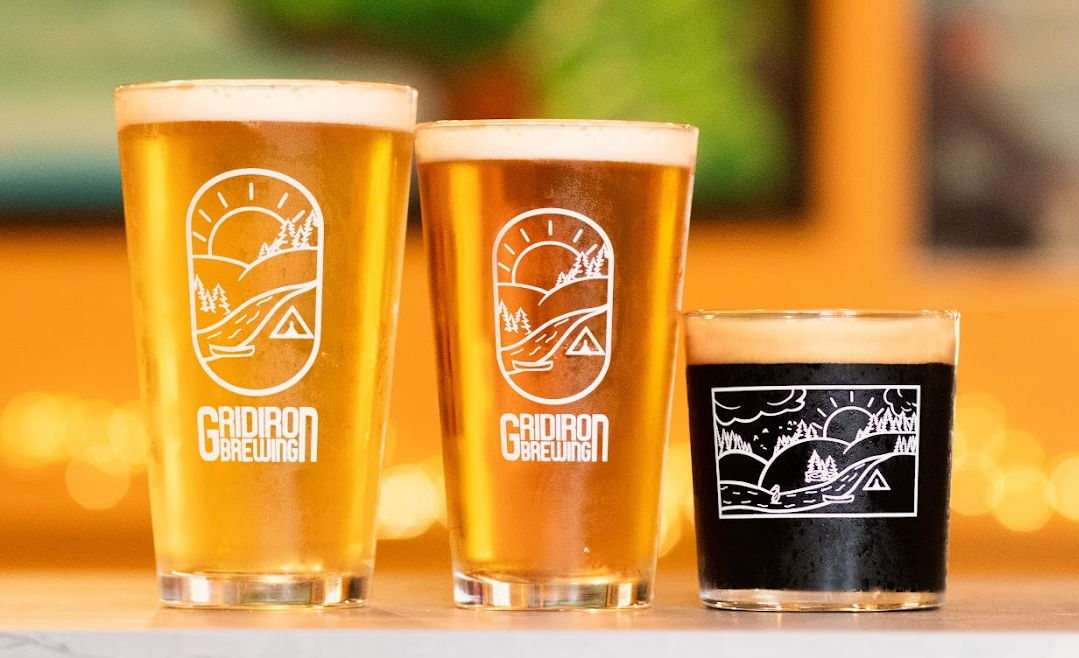 Three glasses of beer, two tall and one short with dark beer, displayed on a wooden surface with a warm blurred background, each featuring a logo of a scenic landscape with a sunrise, trees, and a tent from Gridiron Brewing.