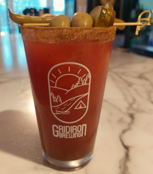 A Bloody Mary cocktail in a glass with a logo of a sunset over mountains and trees, topped with pickles, olives, and a lemon wedge.