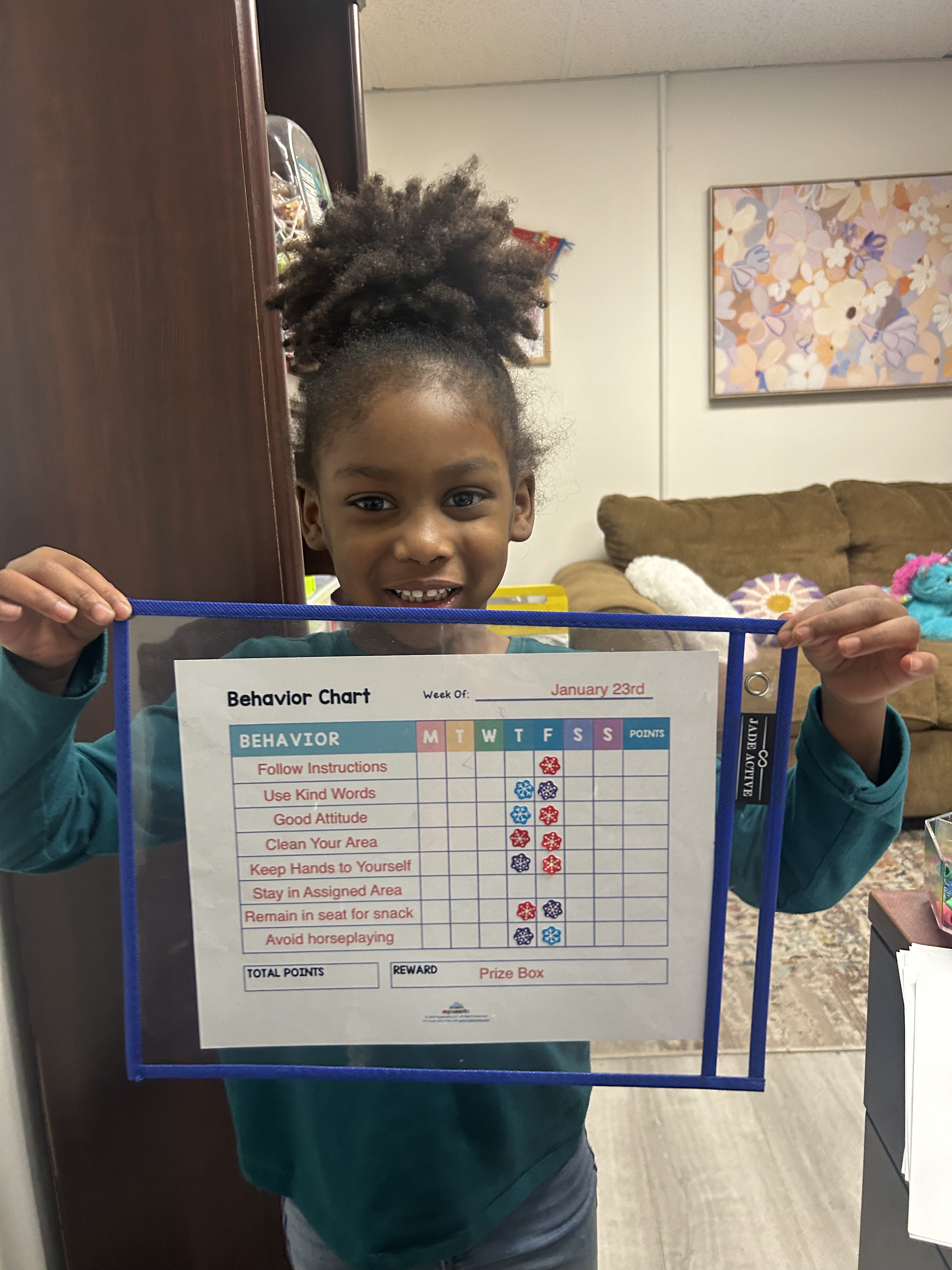 A young girl with curly hair in a ponytail smiling and holding up a behavior chart in a room with a beige couch, colorful abstract wall art, and various plush toys in the background.