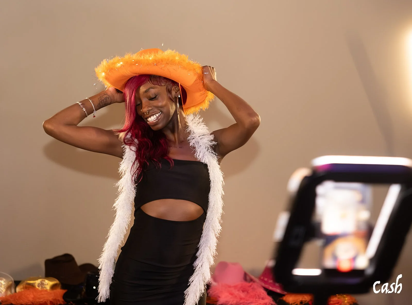 Woman with red hair wearing a large orange carnival hat, a black dress with a cutout, smiling and posing, while being photographed.