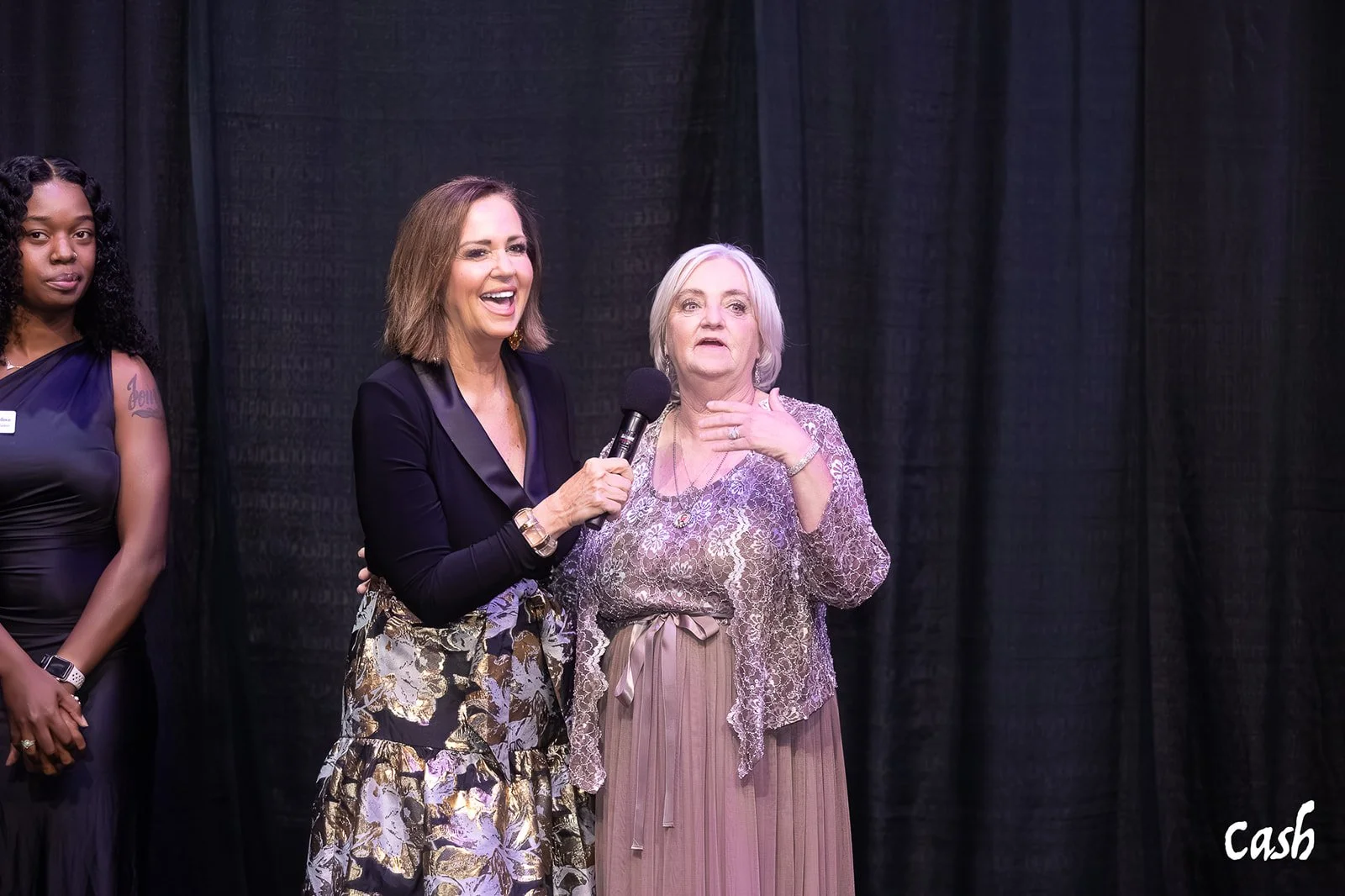 Three women standing on a stage with black curtains. The woman in the middle is holding a microphone and has her hand on her neck, wearing a floral skirt and black top. The woman on the right is wearing a lace top and a tan skirt, looking surprised. 