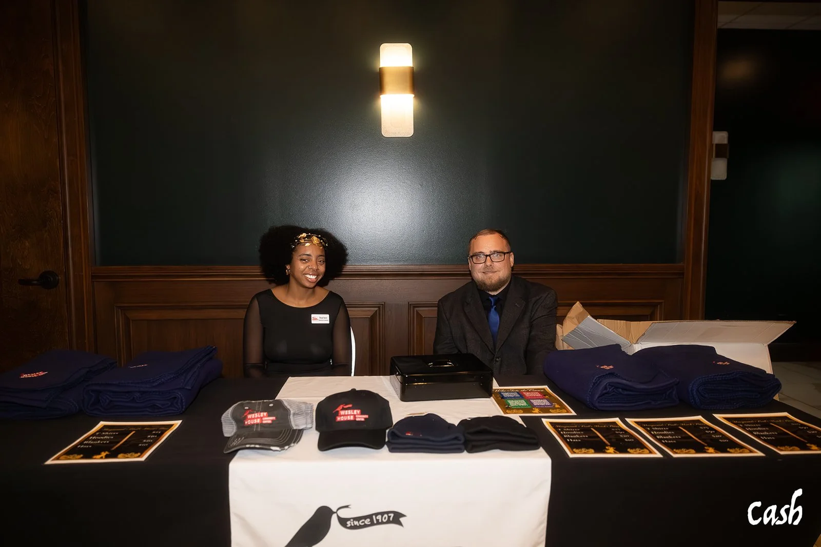 Two people sitting behind a table at a registration or merchandise booth, with various items like caps and shirts displayed, in a dimly lit room with dark green walls.