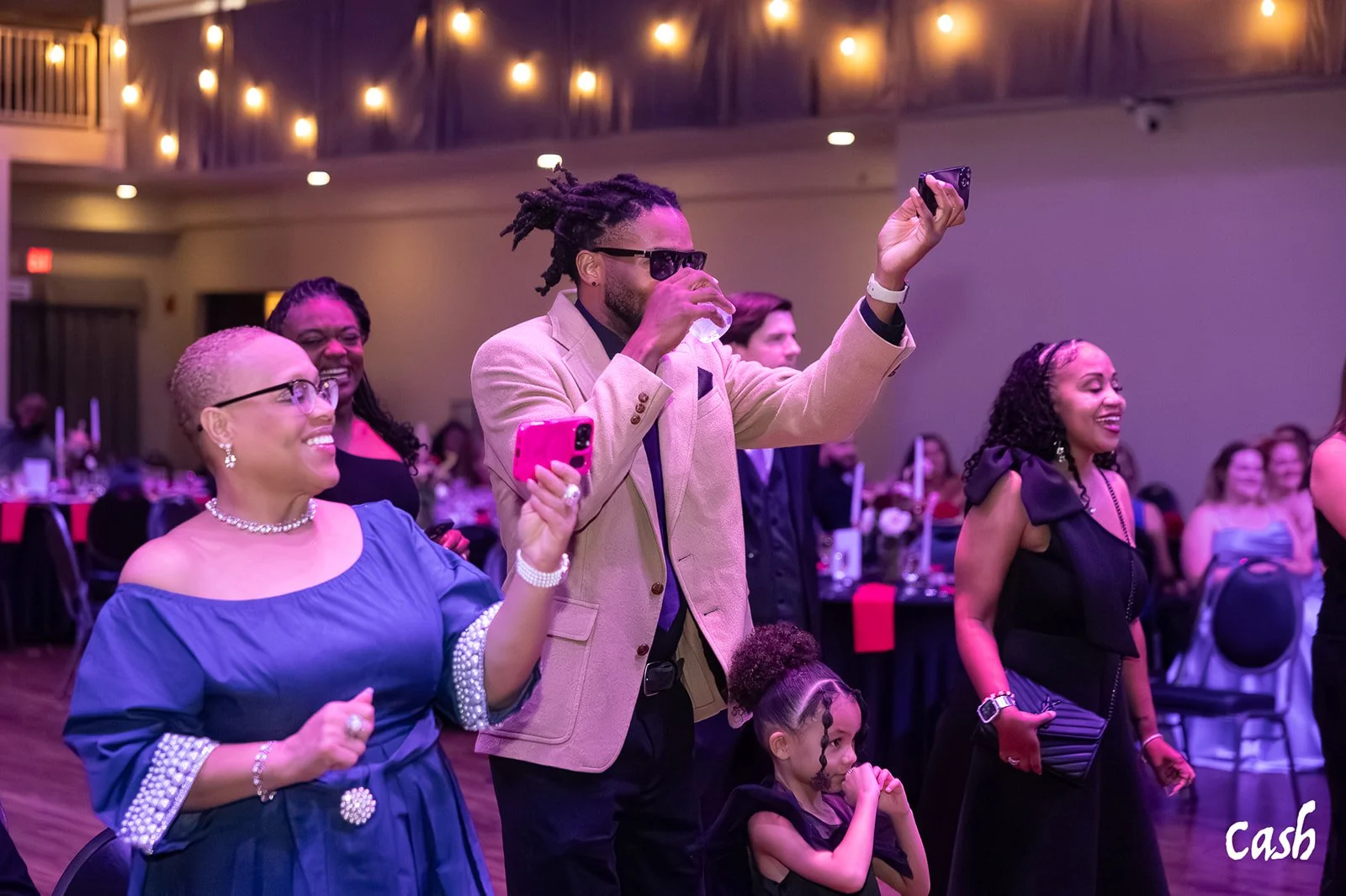 People enjoying a lively event in a decorated venue, with a man taking a selfie with a woman and a child in front, all smiling and dressed elegantly.