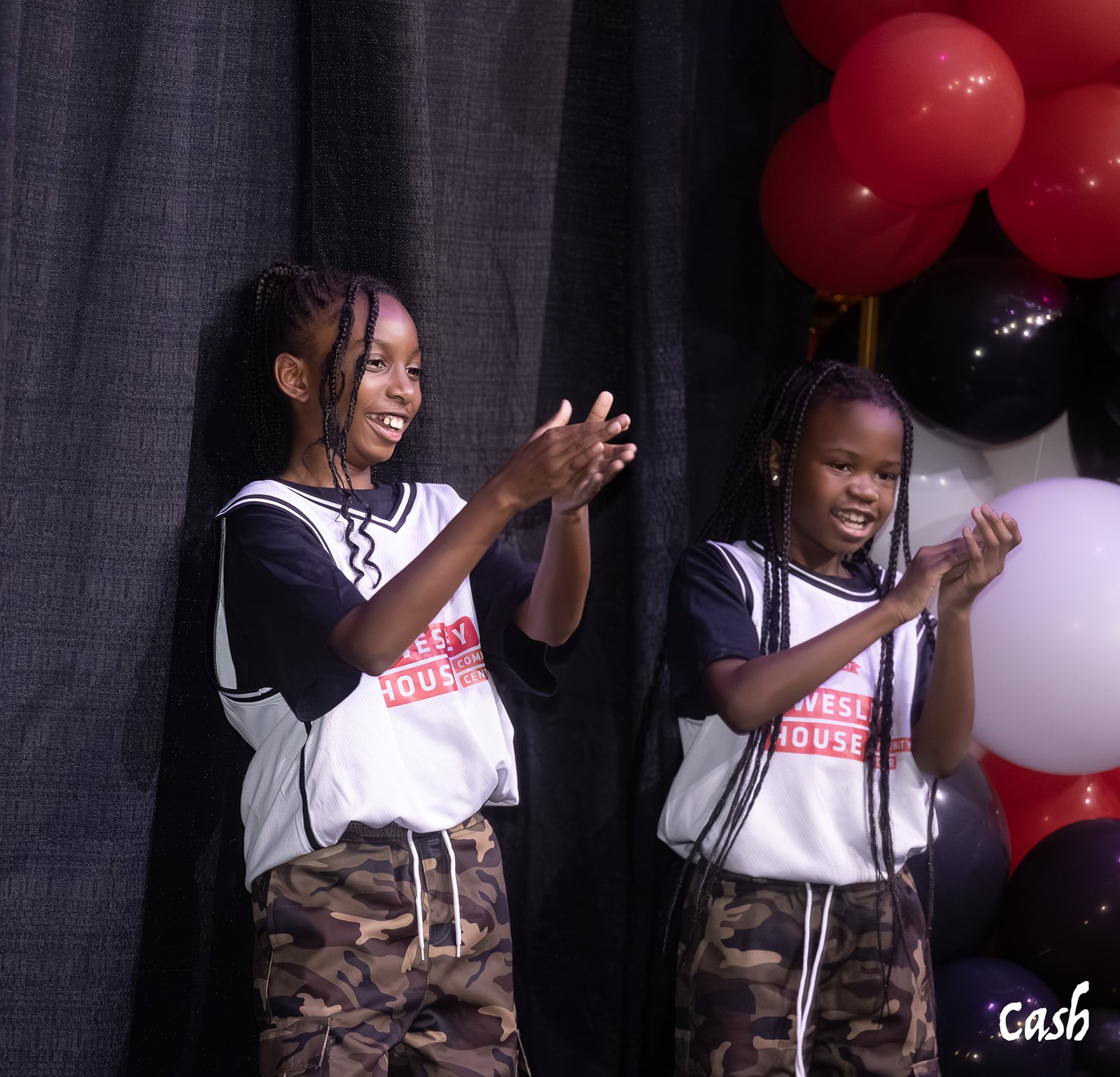 Two young girls with braided hair wearing matching black and white athletic shirts with 'House' printed on them and camouflage pants, standing on stage with black curtains and balloons in black, red, and white in the background, clapping and smiling.