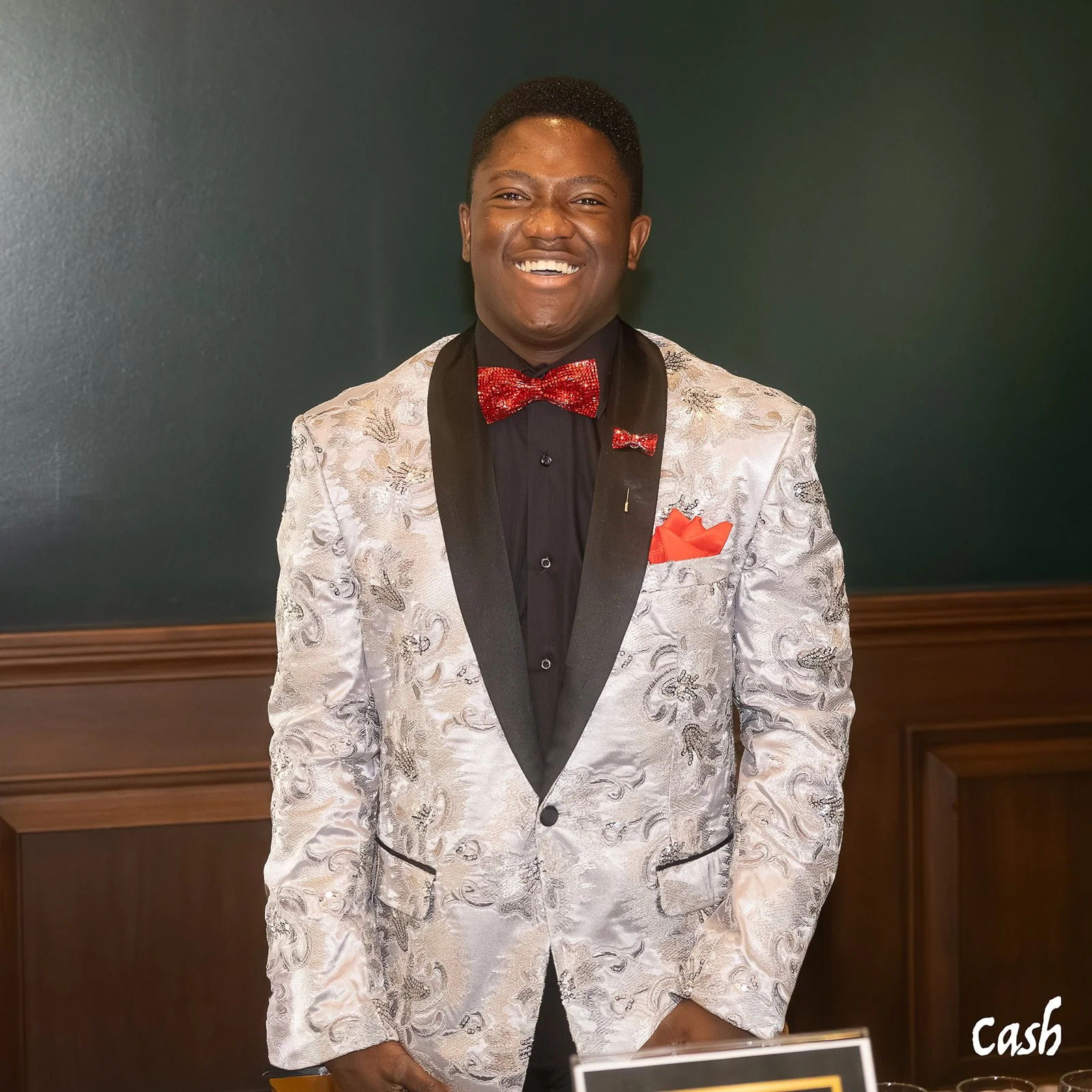 A young man smiling and wearing a shiny silver suit with black lapels, a red bow tie, matching red pocket square, and a black shirt, standing against a dark green wall.