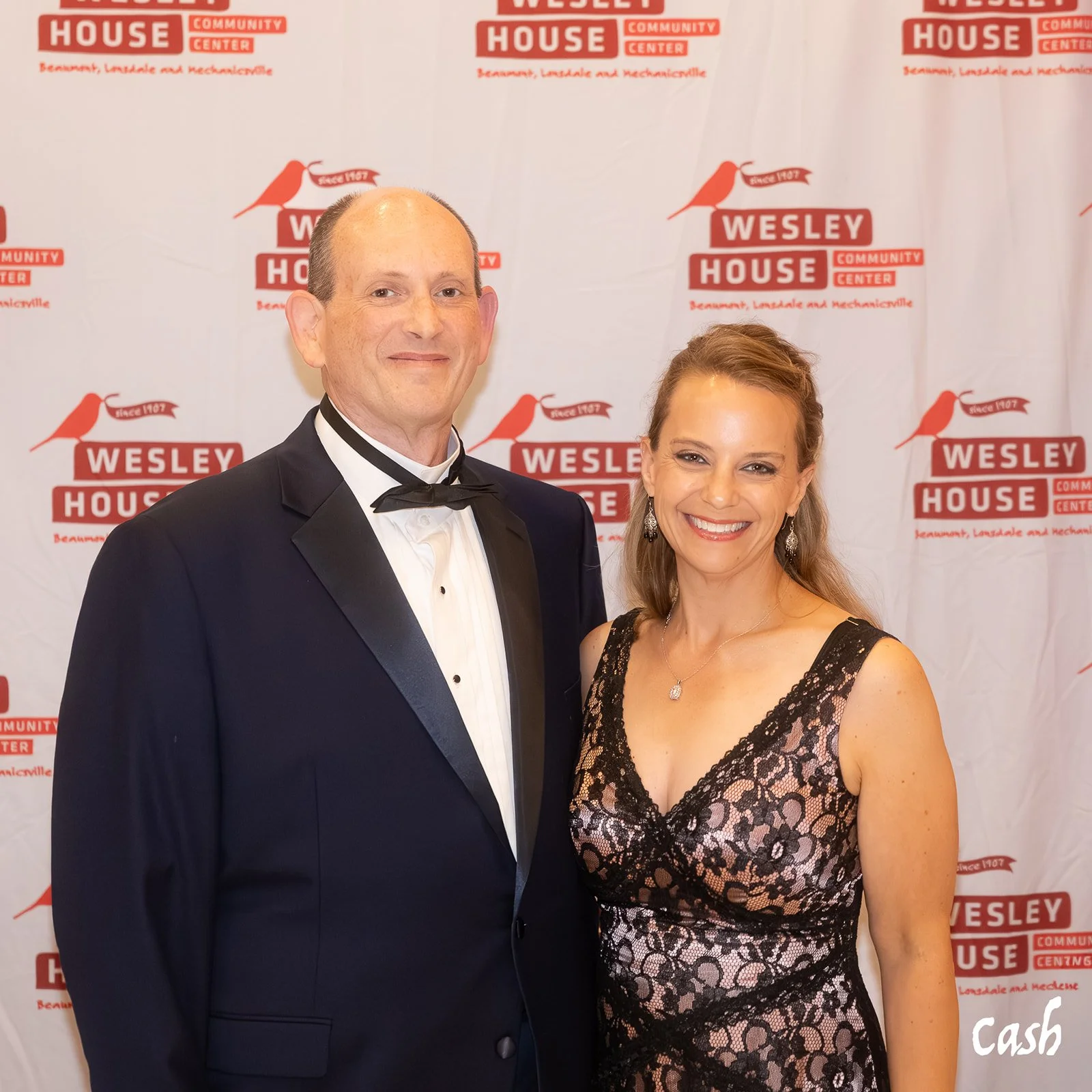 A man in a black tuxedo and a woman in a black and pink lace dress posing together at an event with a Wesley House Community Center backdrop.