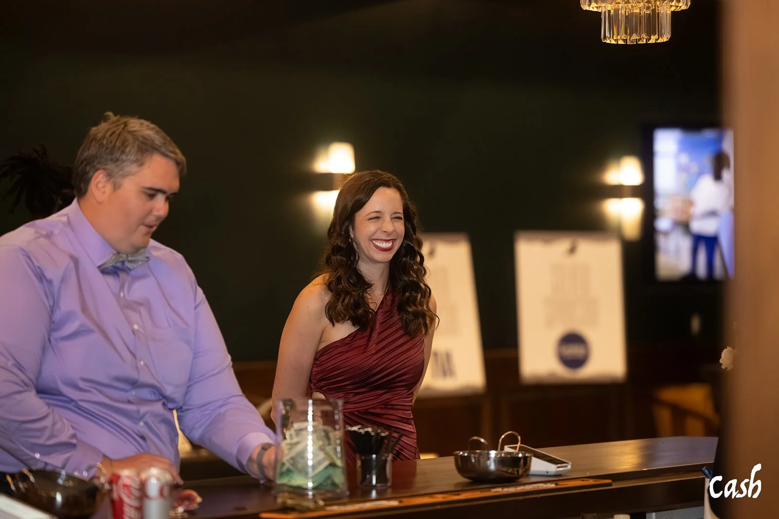 A woman in a one-shoulder maroon dress smiling and laughing at a bar counter, with a male bartender in a purple shirt next to her, in a dimly lit setting.