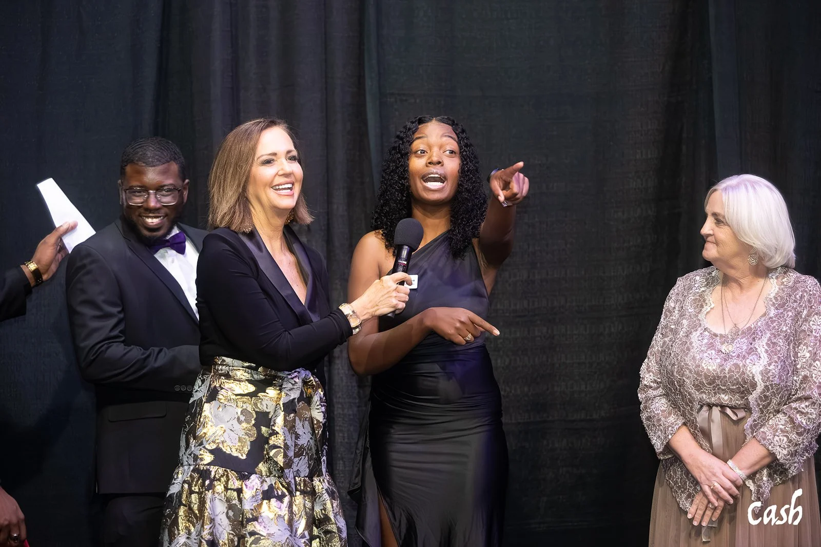Group of five people standing together on stage, with one woman speaking into a microphone and others smiling or looking at her. The scene appears to be at an event or presentation, and there is a dark curtain backdrop. The word 'Cash' is visible in 