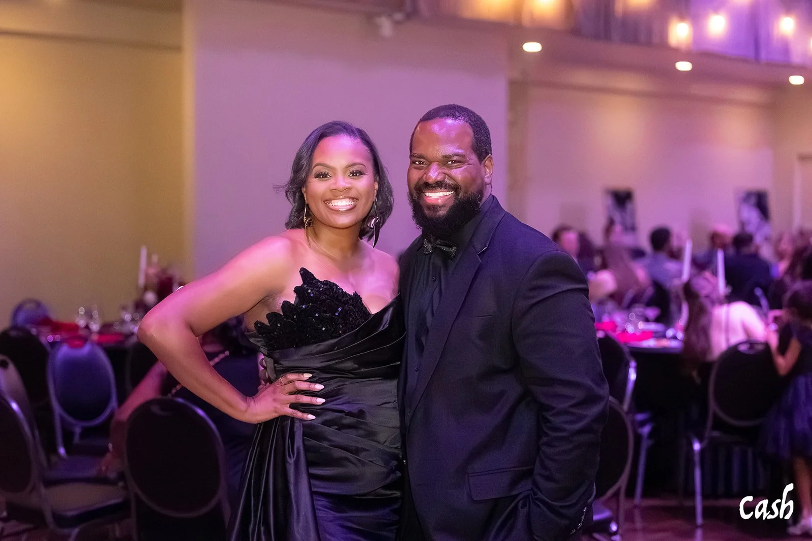 A smiling woman in a black dress with lace details and a man in a black tuxedo and bow tie standing together at a social event or dinner, with tables and other guests in the background.