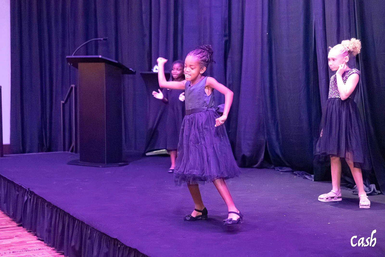 Three young girls in black dresses perform on stage, one dancing with a joyful expression, while the other two observe. The stage has black curtains and a podium.