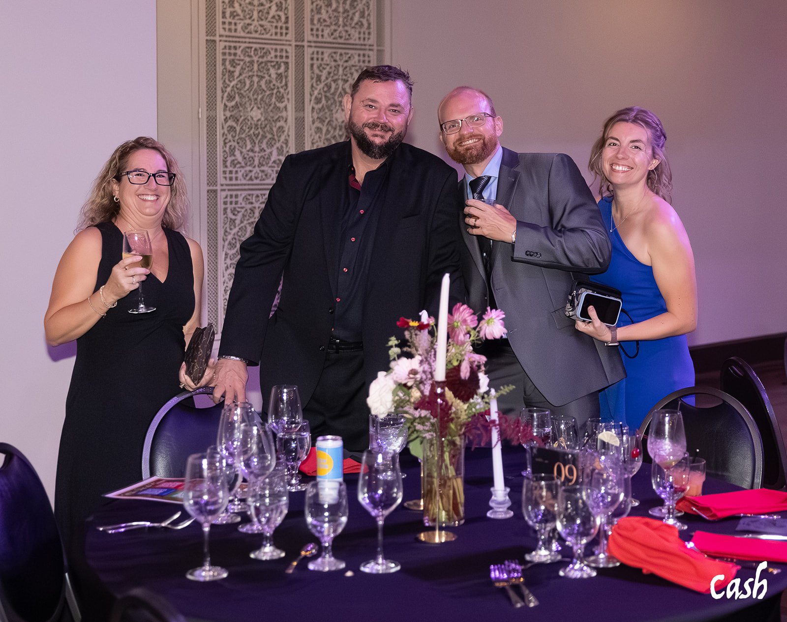 Four adults smiling and posing for a photo at a formal event, standing around a decorated table with flowers, candlesticks, and glassware.