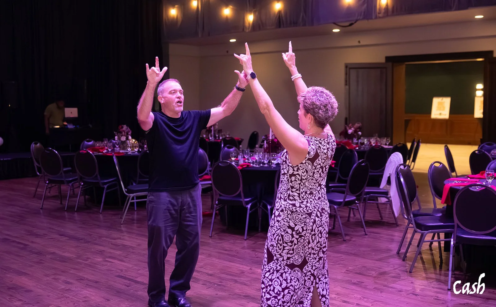 A man and woman dancing together at a celebration, with the man wearing a black T-shirt and gray pants, and the woman in a patterned dress, in a decorated banquet hall with tables and chairs.