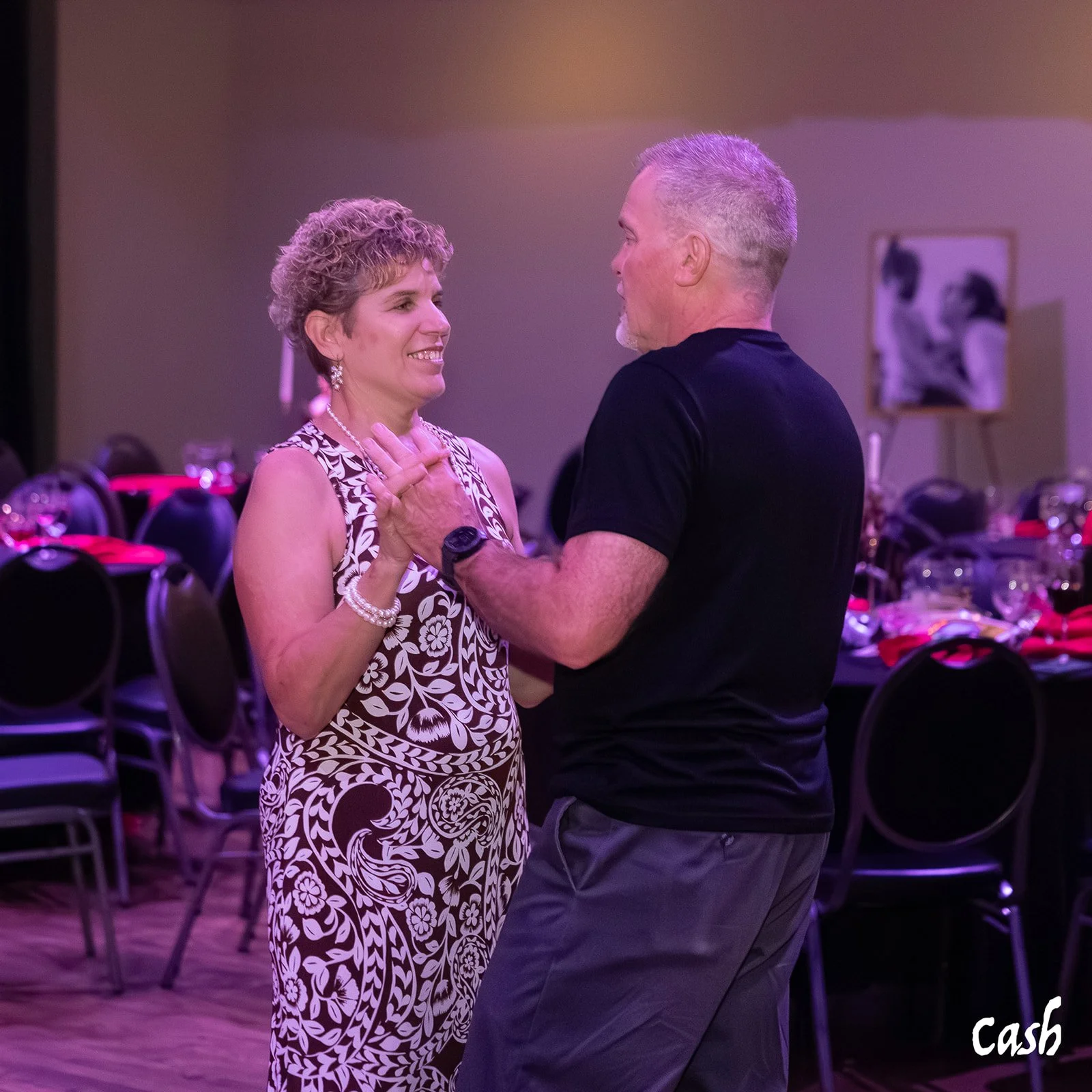 A man and woman dance together at a social event, smiling at each other, in a decorated indoor setting with tables and chairs in the background.