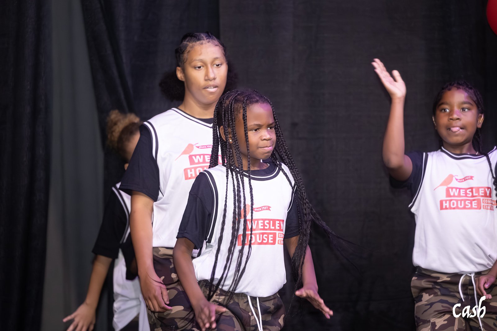 Three young girls performing on stage wearing matching white and black Wesley House t-shirts and camouflage pants, with a black background.