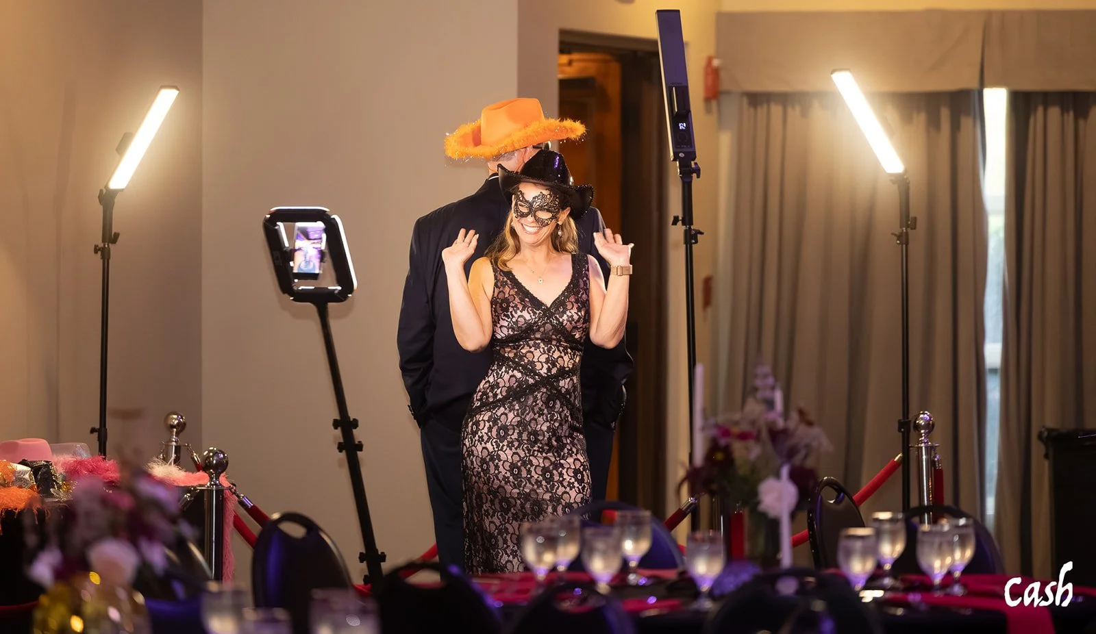 A woman dressed in a black lace dress is smiling and dancing while wearing a black hat and mask. Behind her, a man in a dark suit and an orange hat stands. The scene is set for a photo or video shoot with studio lights and a phone on a tripod capturi