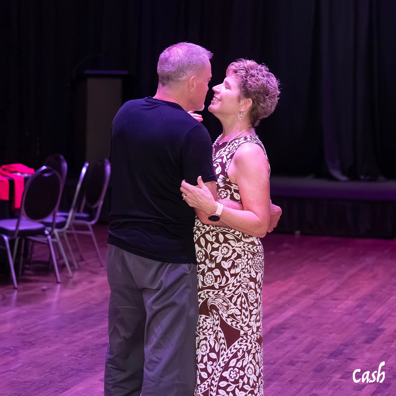 A man and woman dancing closely in a dimly lit room with a black curtain background and empty chairs on the side.