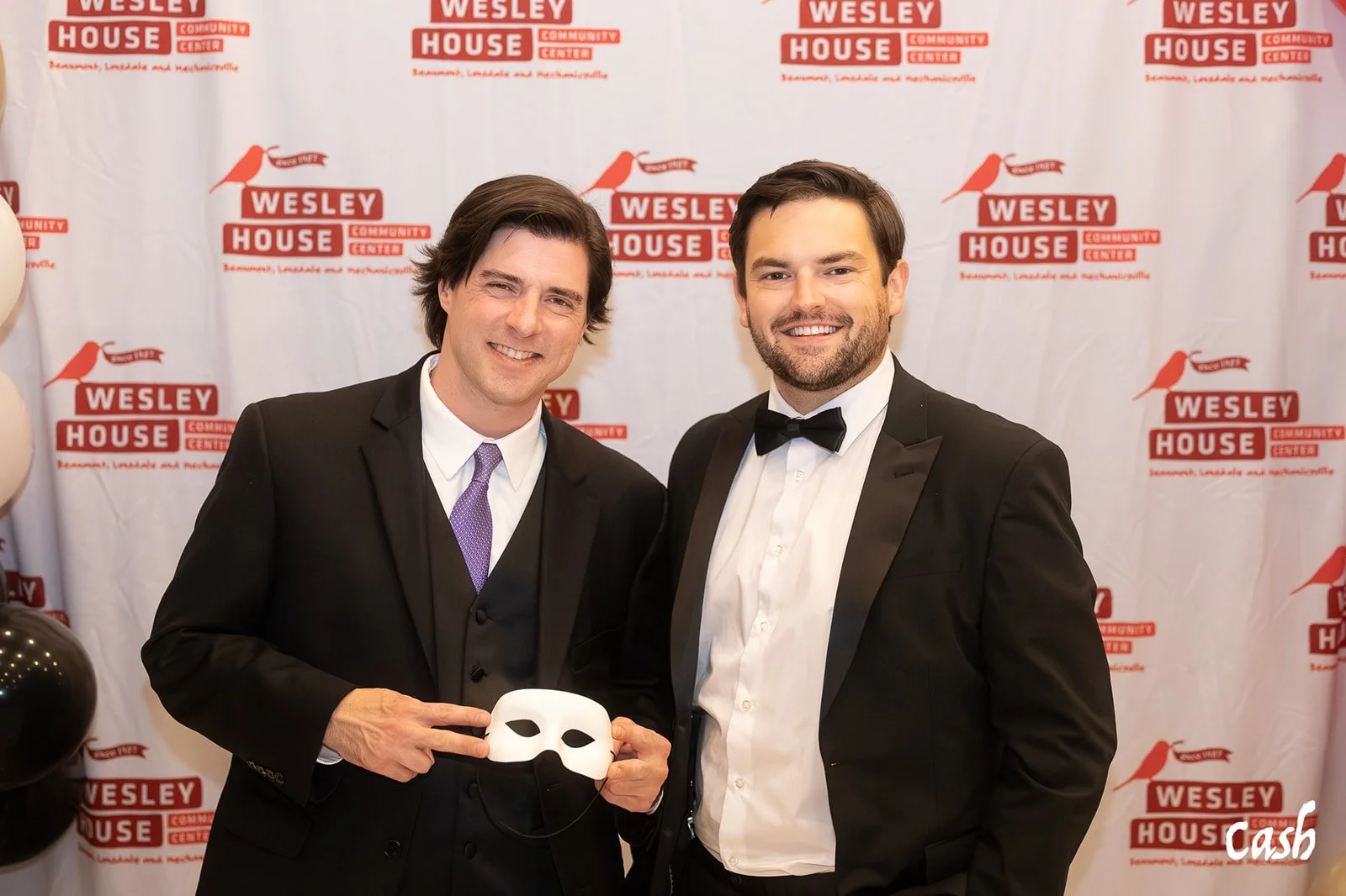 Two men in tuxedos smiling at a formal event with a backdrop that reads 'Wesley House Community Center' and features a bird logo, one holding a white mask and making a peace sign.