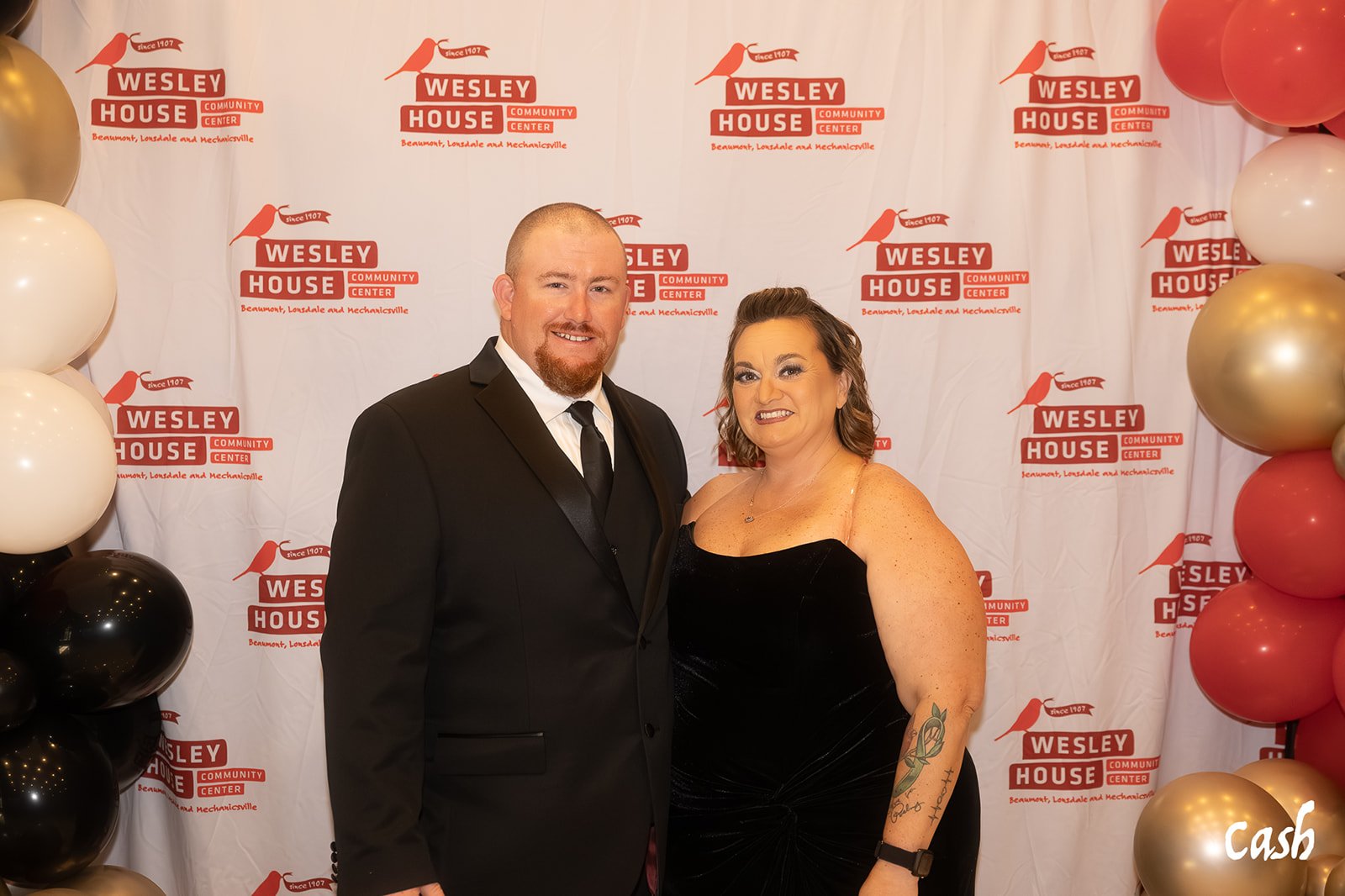 A man in a black tuxedo and a woman in a black dress standing together at an event with a backdrop that reads 'Wesley House Community Center' surrounded by balloons in black, white, gray, pink, and gold.