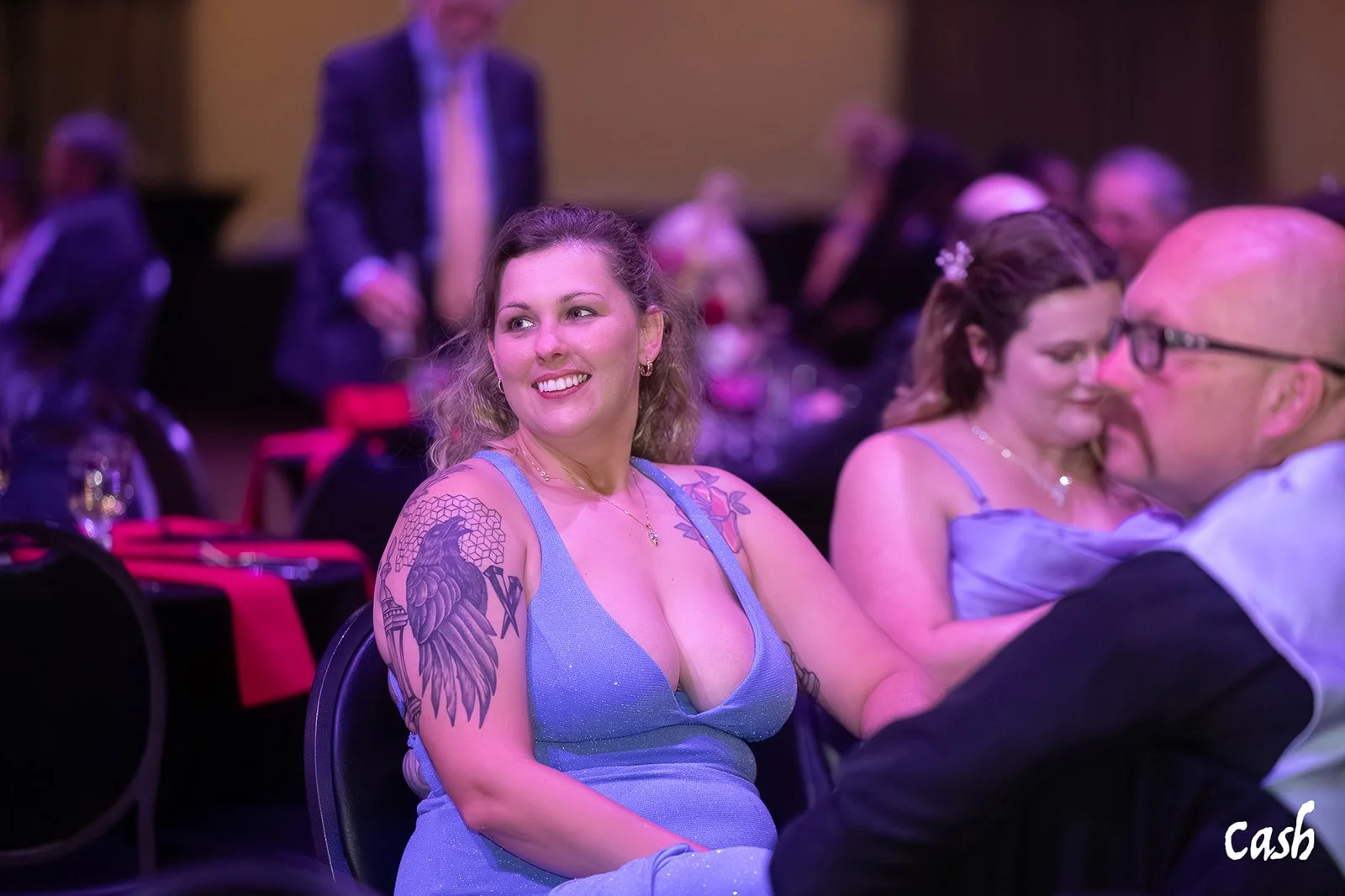 A woman with shoulder-length blonde hair and tattoos on her right arm, wearing a blue dress, sitting at a formal event and smiling. Other people are seated at tables in the background.