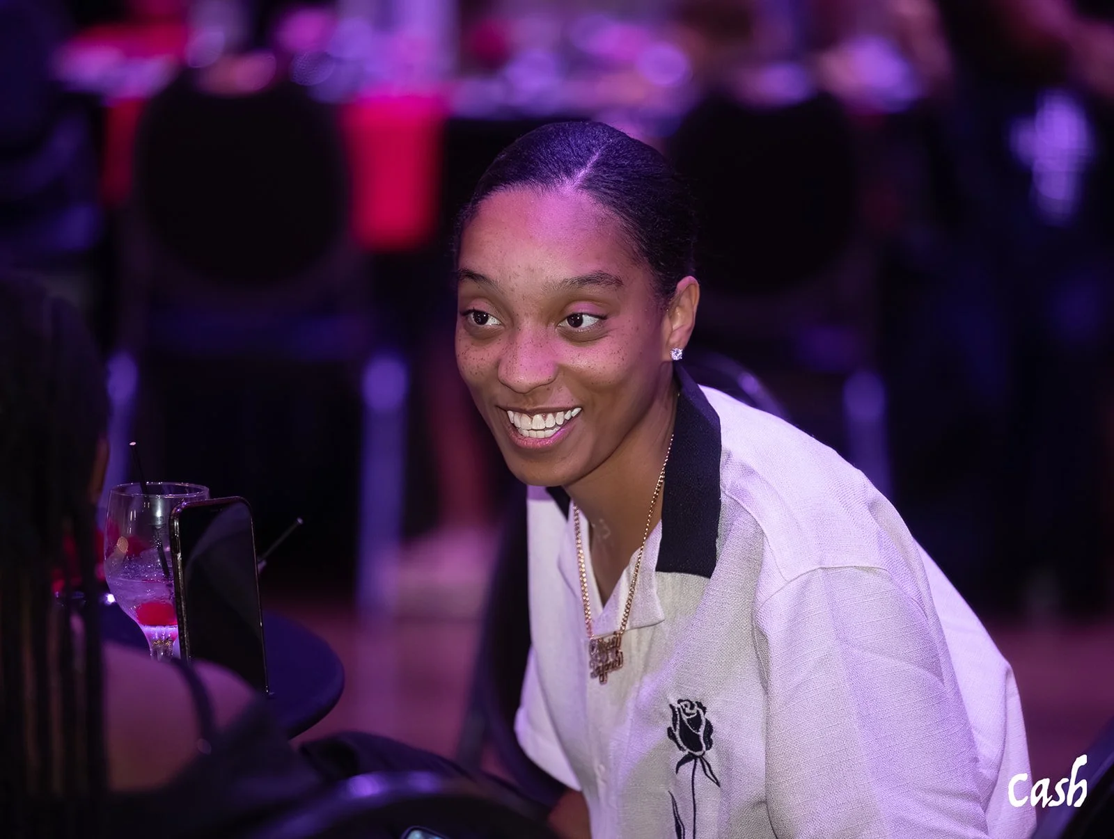 A young woman with dark hair styled in two buns, smiling and engaging in conversation at a social event with purple lighting. She is wearing a beige shirt with a black collar and a rose embroidery, a gold chain, and earrings. There are drinks and a p