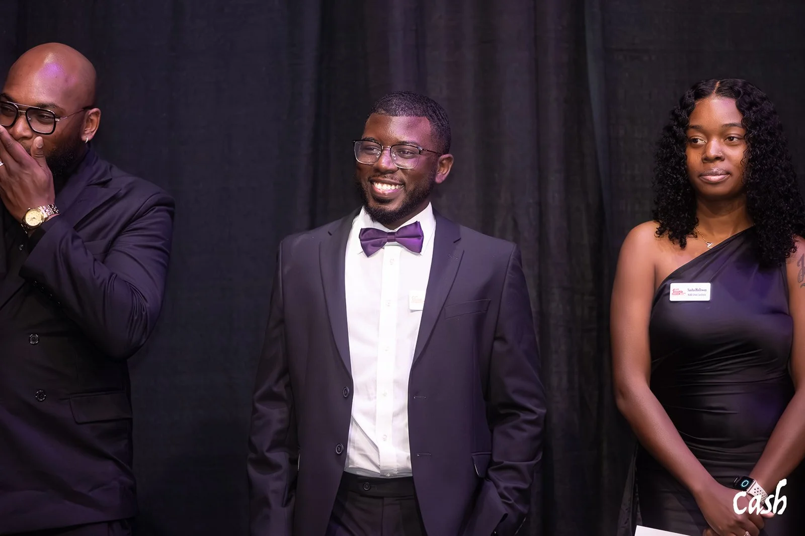 Three well-dressed people standing against a black backdrop, two men and one woman, attending a formal event.