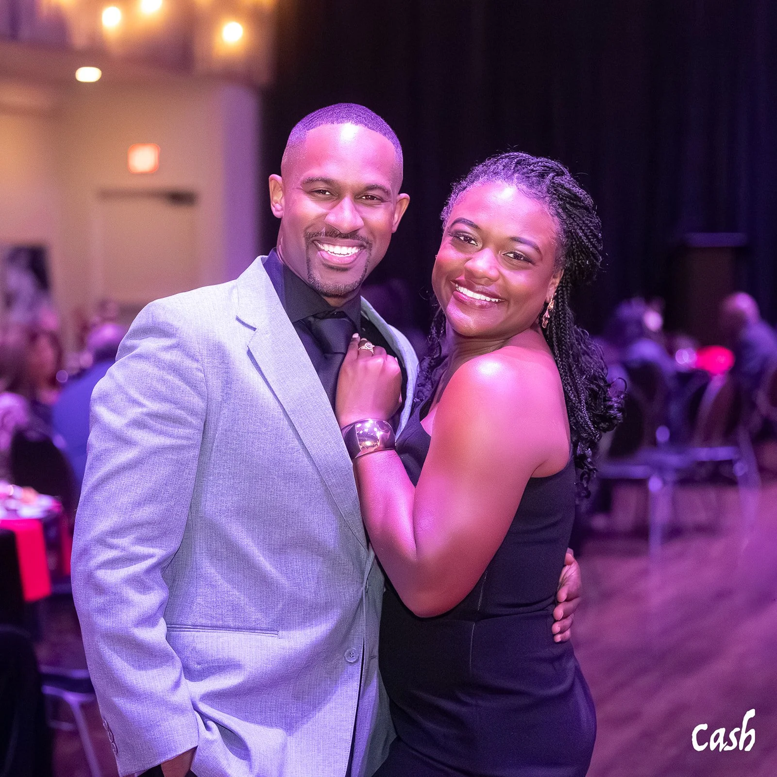 A smiling man in a light gray suit and a woman in a black dress posing together at an indoor event with blurred people in the background.