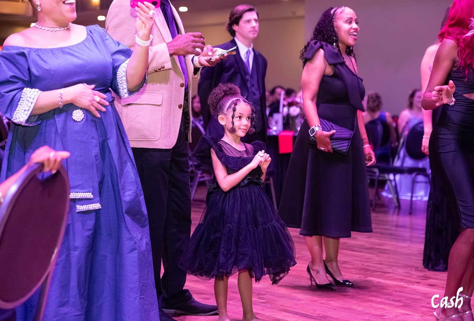 A group of people, including a young girl in a black dress, standing and watching an event at a formal gathering or celebration. The scene is indoors with round tables and chairs in the background.