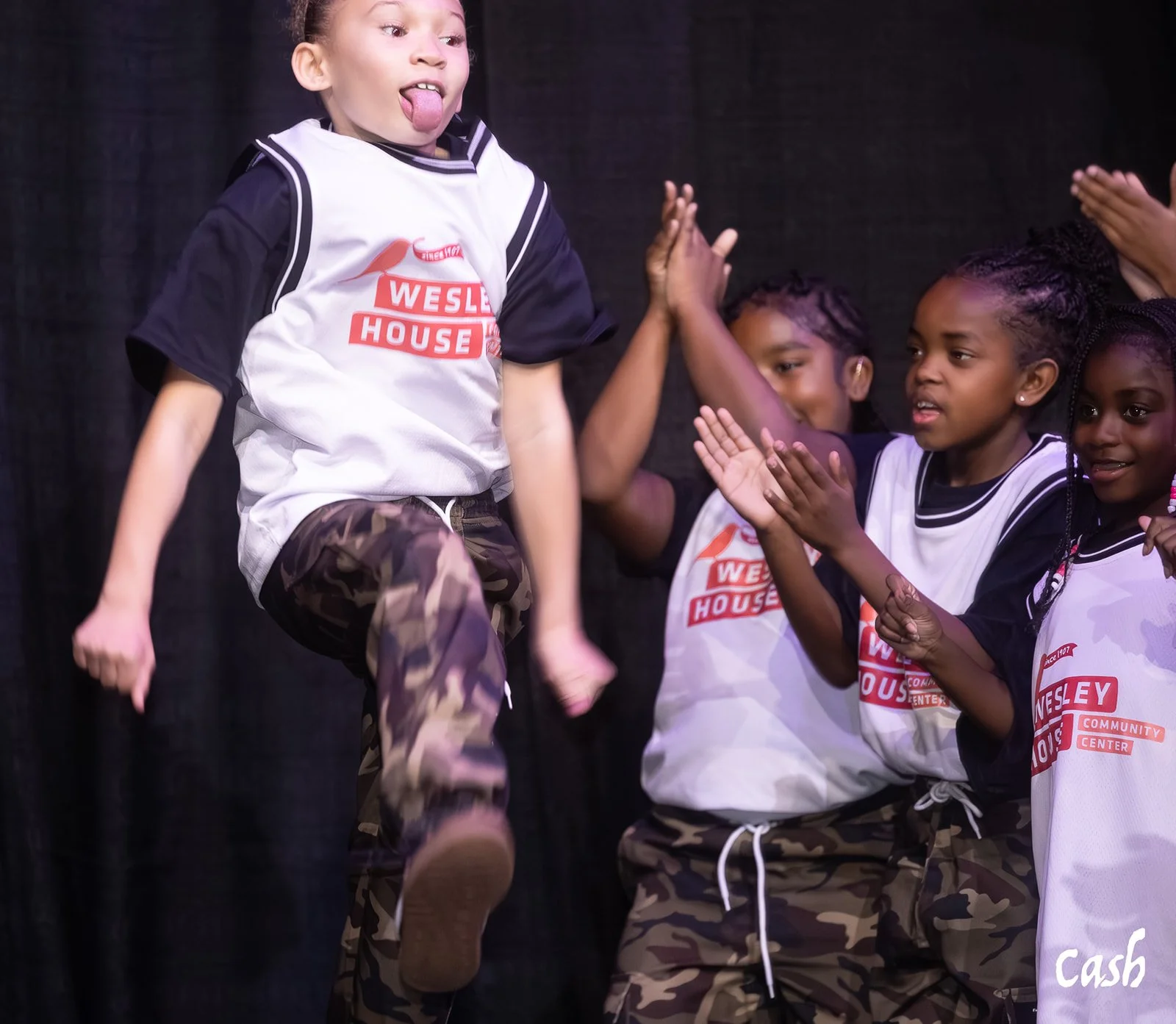 A young girl jumping in front of a group of children giving high fives during a performance on stage.