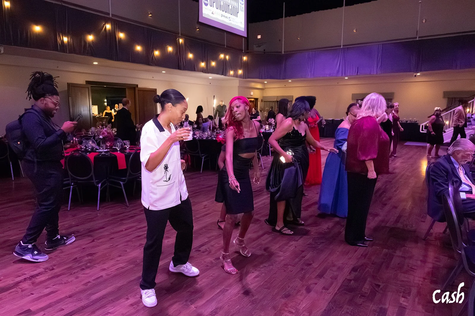 People dancing and socializing at a formal event in a banquet hall with tables, floral centerpieces, and string lights, with a man sitting and looking at his phone on the right.
