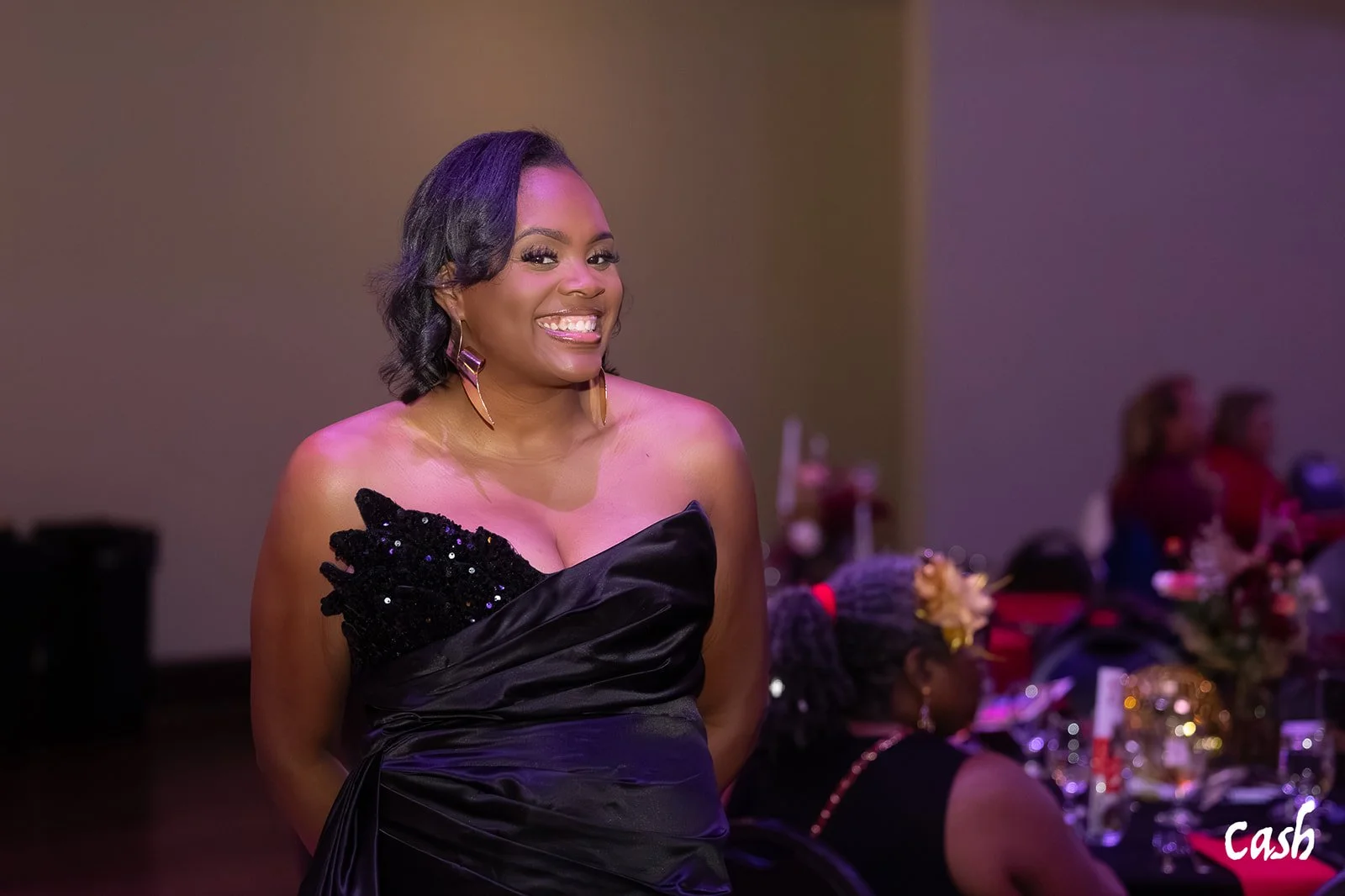A woman smiling, wearing a black strapless dress with sequins, and earrings, at a formal event with tables and other guests in the background.