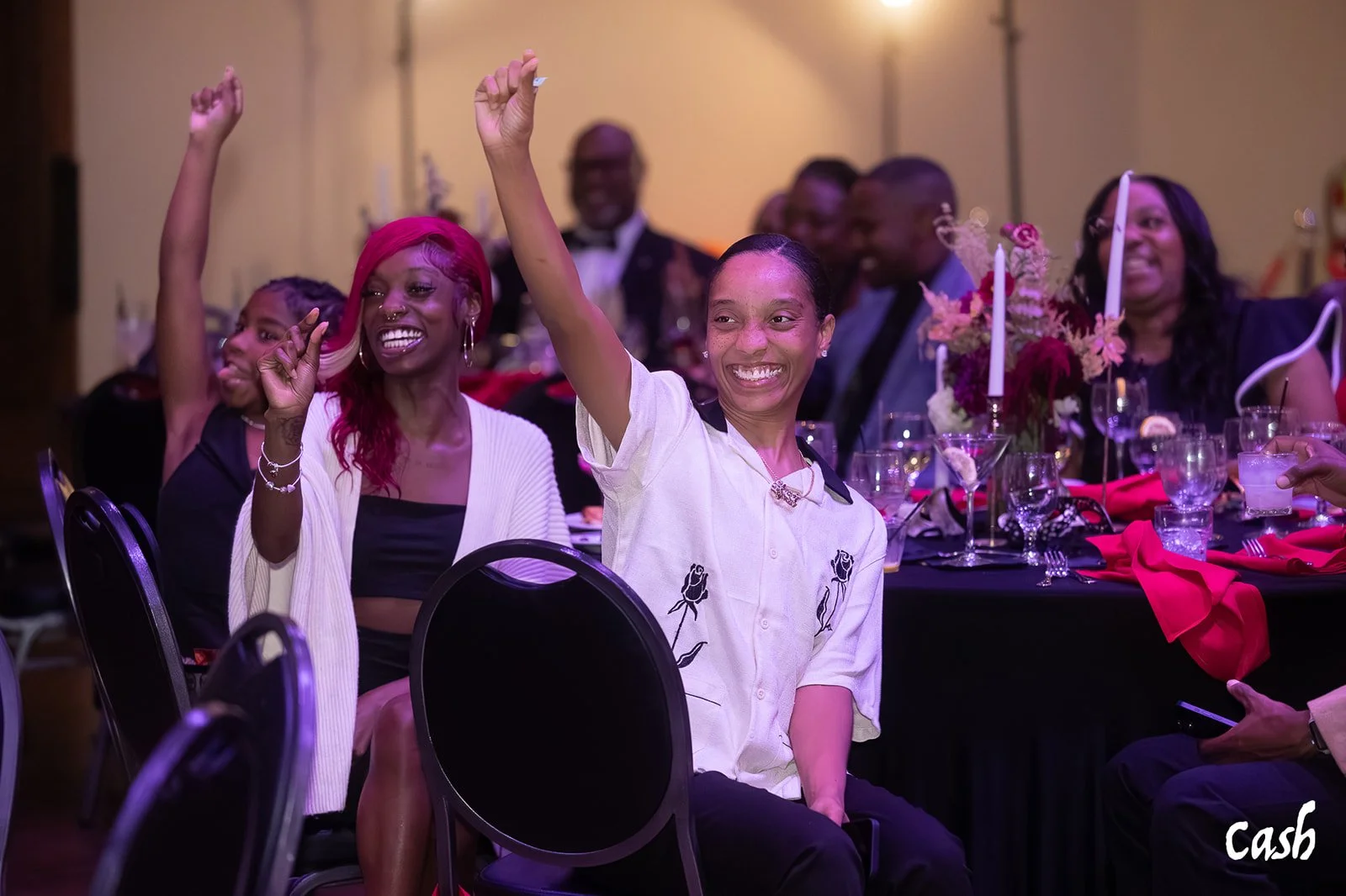 Group of people sitting at a table during an event, smiling and raising their hands, with flowers and candles on the table.