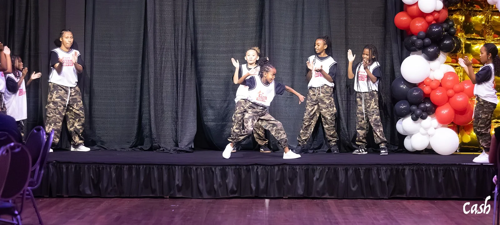 Group of young girls performing a dance on a stage with black curtains, decorated with black, white, red, and gold balloons.