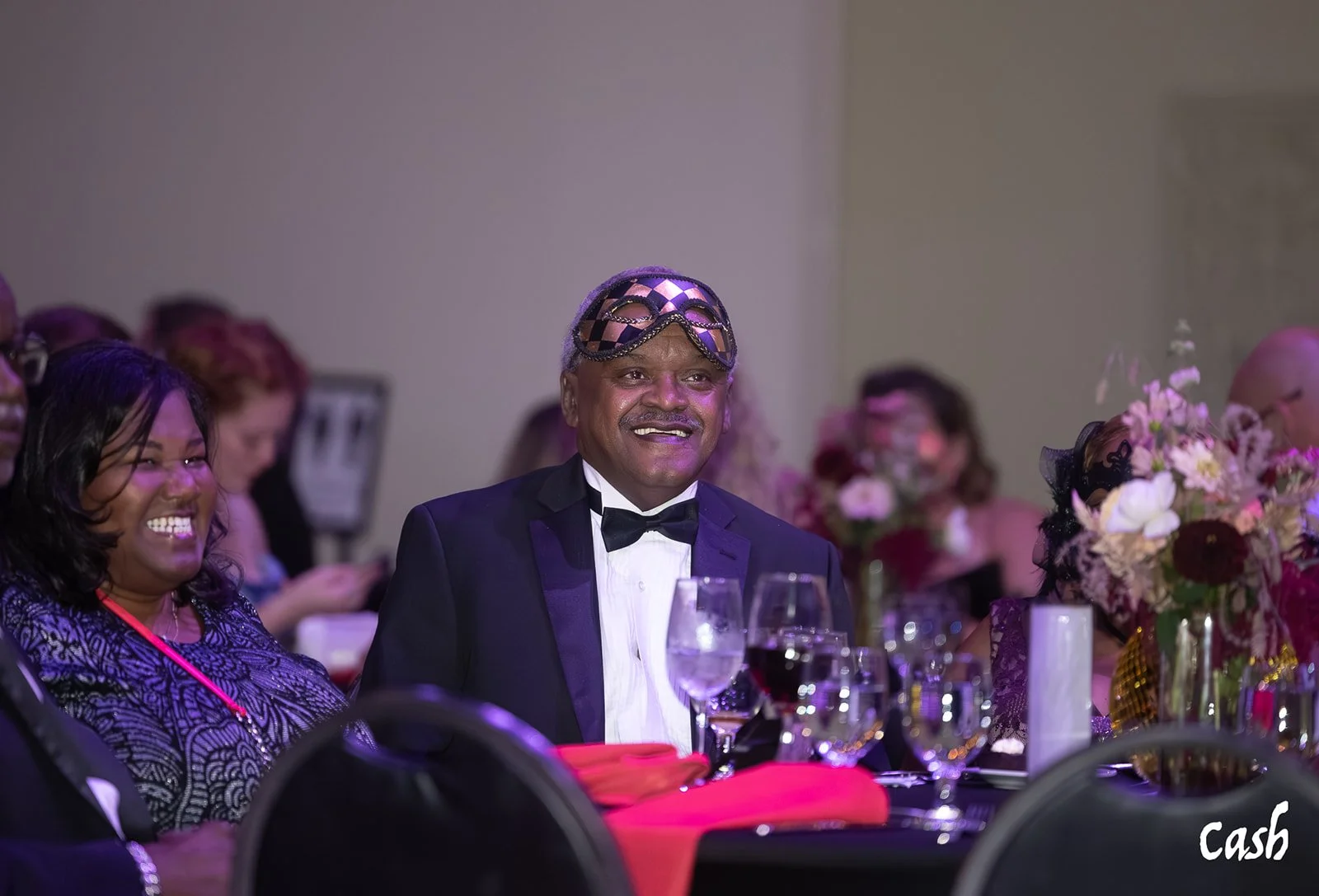 A man wearing a tuxedo and a masquerade mask, sitting at a banquet table surrounded by women and floral centerpieces, smiling during a formal event.