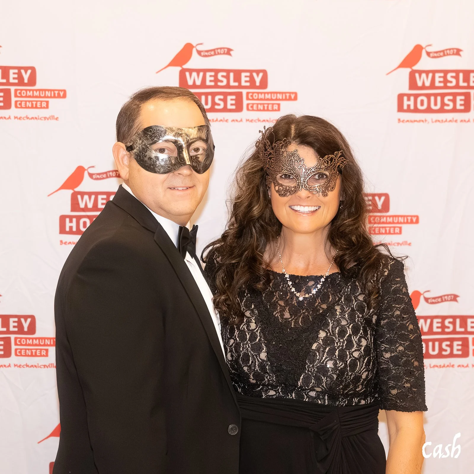 A man and woman in formal attire wearing decorative masks, standing in front of a backdrop for the Wesley House Community Center. The man is in a black tuxedo with a black mask, and the woman is in a black lace dress with a lace mask, smiling at the 